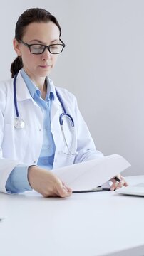 Middle aged female doctor completing healthcare paperwork, clipboard positioned near laptop and stethoscope in well lit clinical workspace by window. Medicine and health care