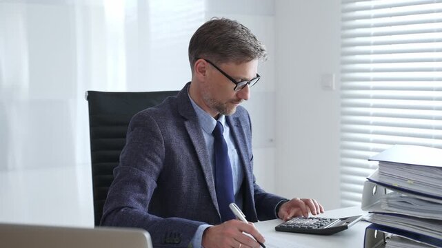 Mature entrepreneur in suit and tie wearing black glasses, is sitting at desk, writing notes and using calculator in modern, bright office. Audit and taxes in business