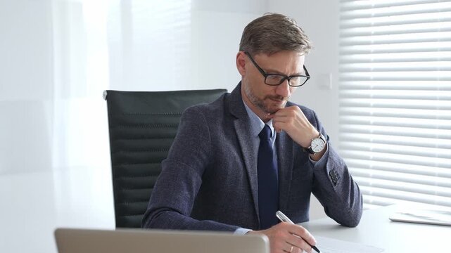 Adult businessman wearing blue suit and tail is concentrating and thinking while working at the desk in a bright, modern office, demonstrating professionalism and efficiency. Business people concept