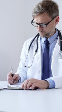 Middle aged doctor man writing a prescription and filling out medical records on a clipboard in an office setting, showcasing the essential aspects of healthcare and patient care. Medicine and health