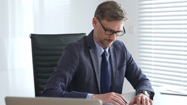 Adult businessman wearing blue suit and tail is concentrating while writing on a document at his desk in a bright, modern office, demonstrating professionalism and efficiency. Business people concept