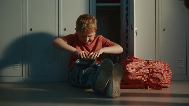 Primary School Boy Sitting on Hallway Floor beside Metal Lockers, Enjoying Burger from Lunch Container During School Break. Childhood, Nutrition, Education, and Everyday Student Life Concept.