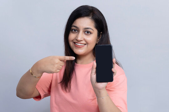 Portrait of an indian smiling young woman holding a smartphone with a blank screen and pointing toward the display, representing mobile technology, digital communication on grey background.