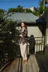 Woman in plaid shirt and light trousers stands on a balcony, natural sunlight highlighting a casual, relaxed lifestyle and urban mood, with warm tones and a tranquil, everyday fashion feel © SHOTPRIME STUDIO