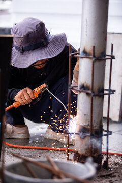 Selective focus to Male construction worker wearing sunglasses and bucket hat welding a steel structure at a job site. Glowing bright sparks fly off the electric metal arc welding process during outdo