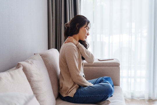Woman suffering from neck pain while sitting on the couch at home, Health care and medical concept