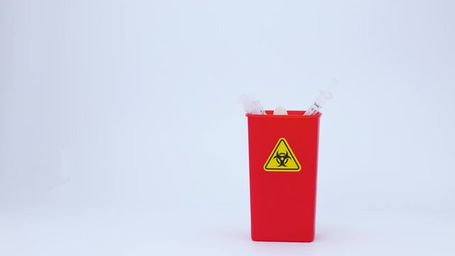 Gloved hand carefully dropping medical syringe needle into red biohazard clinical waste container feeling cautious under studio light