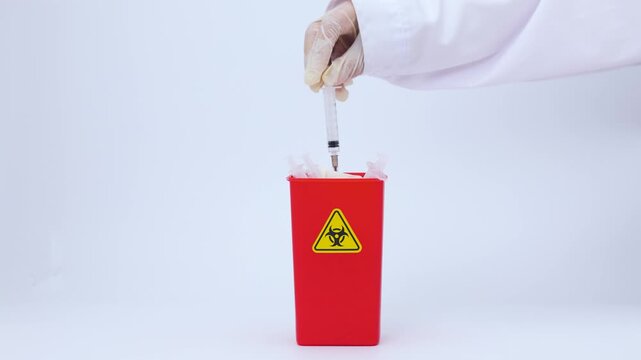 Gloved hand throws syringe clinical waste into red sharps container with biohazard symbol showing cautious professional mood