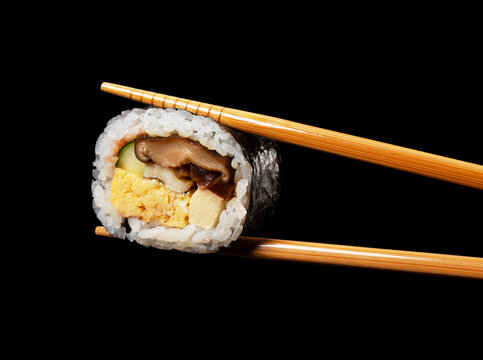 Chopsticks Holding a Piece of Japanese Futomaki Thick Sushi Roll Isolated on Black Background