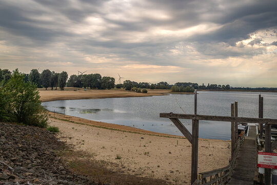 Der See lag ruhig unter dem dramatischen Wolkenhimmel, w&auml;hrend die Sonnenstrahlen wie goldene Finger durch die Wolkendecke griffen. Am Ufer gegen&uuml;ber die Windr&auml;der, der Sandstrand und die weite Landsc