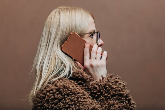 A stylish blonde woman wearing a teddy coat with phone standing against a rich brown background in shade Mocha Mousse