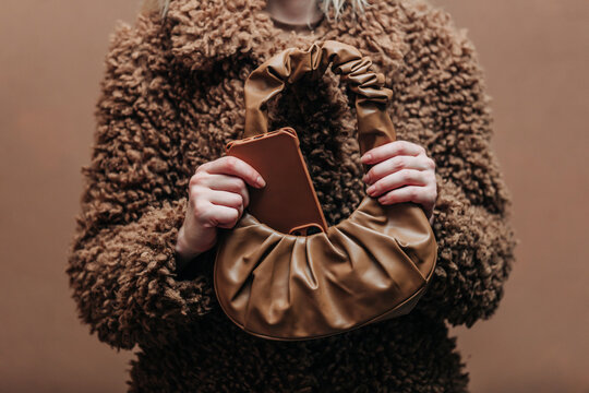 A stylish blonde woman wearing a teddy coat putting phone in a bag and standing against a rich brown background in shade Mocha Mousse