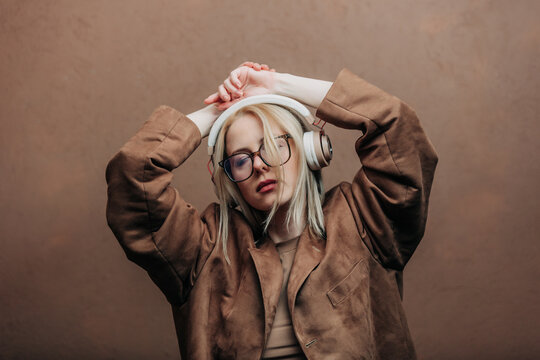 A stylish blonde woman wearing a brown jacket paired with modern headphones, standing against a rich brown background in shade Mocha Mousse