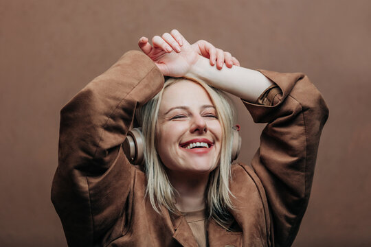 A stylish blonde woman wearing a brown jacket paired with modern headphones, standing against a rich brown background in shade Mocha Mousse
