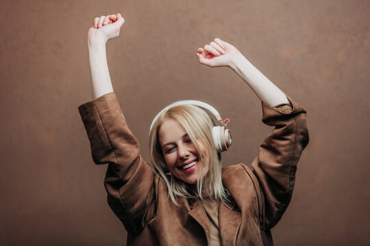A stylish blonde woman wearing a brown jacket paired with modern headphones, standing against a rich brown background in shade Mocha Mousse