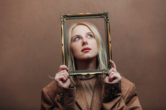 A stylish blonde woman wearing a brown jacket paired with vintage picture frame, standing against a rich brown background in shade Mocha Mousse