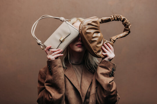 A stylish blonde woman wearing a brown jacket paired with vintage bags, standing against a rich brown background in shade Mocha Mousse