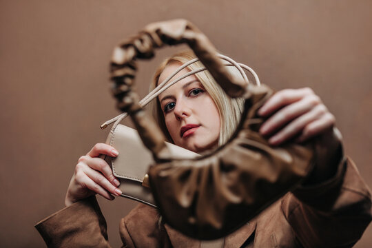 A stylish blonde woman wearing a brown jacket paired with vintage bags, standing against a rich brown background in shade Mocha Mousse