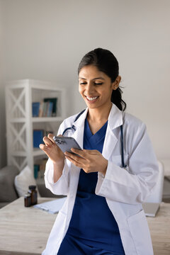 Smiling Indian female doctor standing in workplace using smartphone, reviewing medical information, conducts digital communication, remote consultation with patient via mobile telehealth application