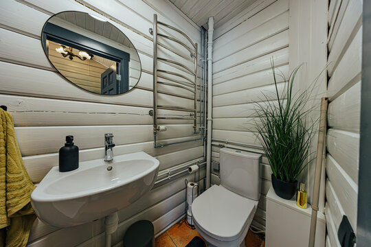 Small bathroom with white-painted timber walls, wall-mounted sink, toilet, oval mirror, heated towel rail, exposed pipes, green plant, and decorative accessories