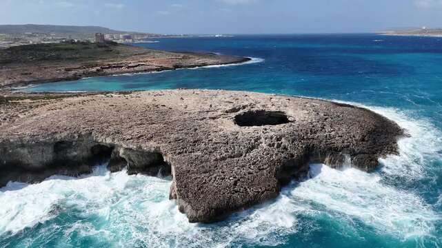 limestone headland coral lagoon malta juts into mediterranean sea turquoise cove waves crash white surf sea caves undercut cliff base distant coastal town skyline hazy horizon aerial establishing shot