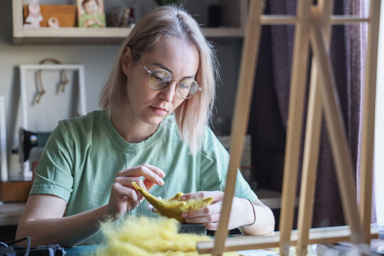 Young Woman With Glasses Carefully Needle Felts Yellow Wool Figure At Home Craft Table, Working With Focused Expression In Soft Natural Window Light Inside Cozy Creative, With Felting, Needle, Wool