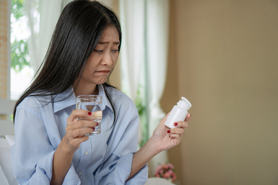 Unhappy young Asian woman holding a glass of water and a white pill bottle, looking disgusted while sitting on a bed. Concept of illness, reluctance to take medicine, and healthcare.