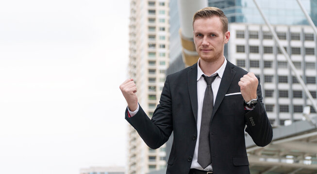 Confident Caucasian businessman in black suit making fist gesture of success and victory while standing outdoors in modern city. Corporate achievement, determination and business motivation concept.