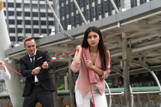 Determined Asian businesswoman and Caucasian businessman in suit pulling red rope together in a modern city street. Business competition, teamwork, and corporate struggle concept.