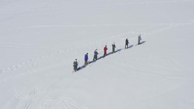A group of people wearing traditional costumes cross country ski together over vast snow covered field on bright sunny day.