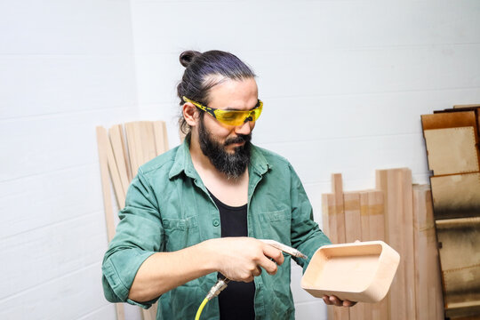 Bearded Craftsman In Green Work Shirt Uses Spray Tool To Apply Finish To Unfinished Wooden Tray Inside Bright Workshop, Wearing Yellow Safety Glasses And Working Near Stacked Raw Wood