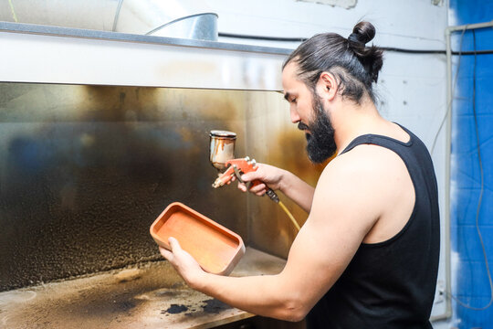 Bearded Man In Black Tank Top Spray Coats Wooden Tray Inside Industrial Workshop, Using Handheld Spray Gun With Focused Precision While Working Beside Large Metal Finishing Chamber