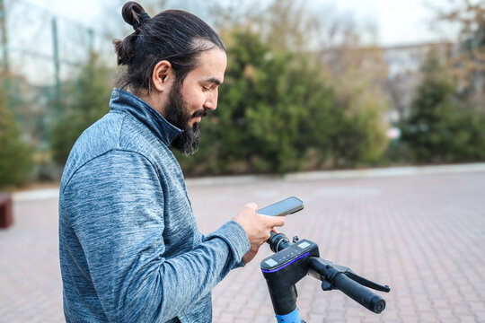 Bearded Man In Blue Jacket Stands Beside Electric Scooter While Checking Smartphone On Paved Outdoor Path, With Shallow Depth Of Field, Soft Daylight, Blurred Trees, And Relaxed Everyday City Travel