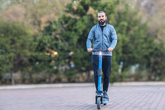 Bearded Man Rides Electric Scooter Along Paved Park Path, Facing Forward With Relaxed Expression, Blue Jacket, Dark Pants, And Soft Green Bokeh Background Suggesting Modern Urban Mobility
