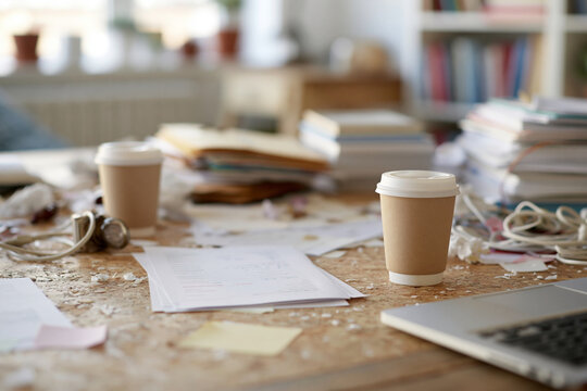 Chaotic desk shows coffee cups, papers, wires, and books. Laptop rests near scattered notes and tangled cables. Natural light fills the room, suggesting daytime work. Clutter implies busy, creative
