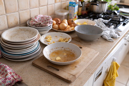 Kitchen counter cluttered after a busy meal. Dishes stacked, bowls empty, utensils nearby. Yellow towel hangs, gloves rest beside stove. Natural light illuminates worn tiles and wood