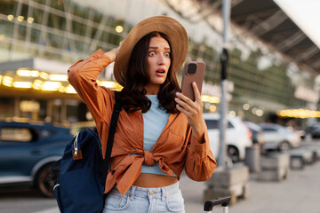 Shocked young woman looking at smartphone while standing with backpack and suitcase outside modern...