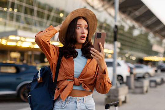 Shocked young woman looking at smartphone while standing with backpack and suitcase outside modern airport terminal