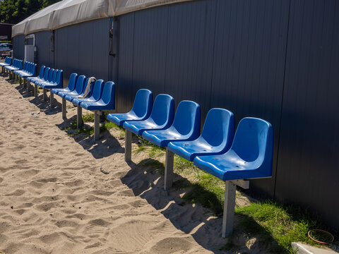 Blue Spectator Seats beside Sandy Beach Soccer Field. A row of bright blue spectator seats stands along the edge of a sandy outdoor sports field. The seating area is positioned beside a beach soccer 