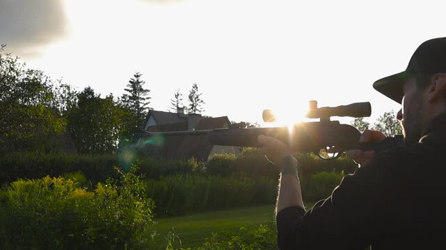 Young man wearing a baseball cap aiming and shooting a rifle equipped with a telescopic sight. Beautiful golden hour sunlight creates a lens flare over the lush green suburban backyard setting