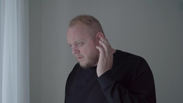 Worried man in a dark sweatshirt pauses and touches his ear, reacting to sudden ringing and possible tinnitus indoors.