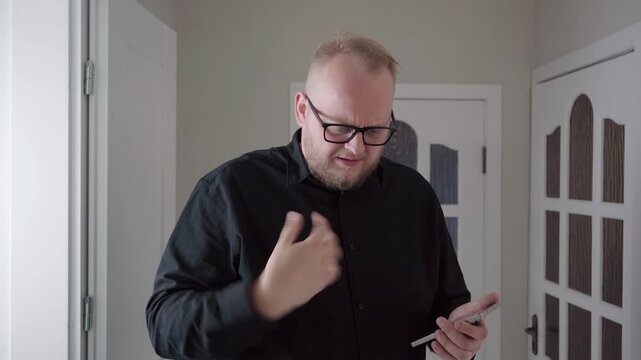 Middle-aged man in glasses scrolls his smartphone and reacts with thumb pain, suggesting overuse or repetitive strain indoors.