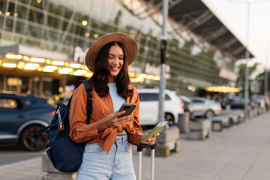 Smiling lady using her smartphone while holding passport, standing with suitcase outside modern airport terminal, free space