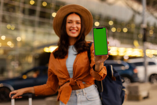 Cheerful lady displaying smartphone screen for advertising or travel app mockup while waiting with luggage outdoors of airport terminal, mockup