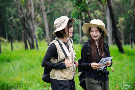 Two happy young Asian female friends hiking together in green forest, carrying backpacks, enjoying eco travel and nature.