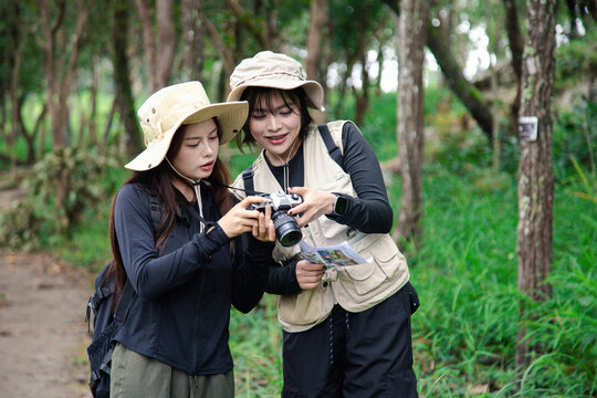Two happy young Asian female friends hiking together in green forest, carrying backpacks, enjoying eco travel and nature.