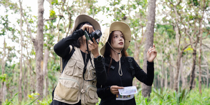 Two happy young Asian female friends hiking together in green forest, carrying backpacks, enjoying eco travel and nature.