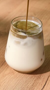Hojicha tea pours into oat milk over ice in clear glass, creating layered iced latte with swirling roasted tea on wood table.