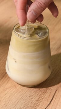 Hand stirs iced hojicha latte in clear glass, mixing roasted tea with oat milk and ice into creamy drink on wooden table.