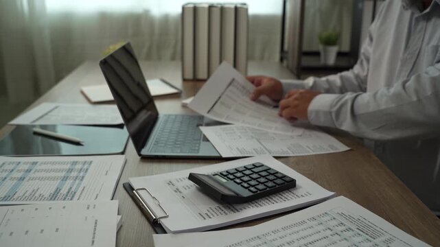 Accountant using calculator while reviewing financial reports with laptop, tablet, pen for accounting, tax preparation, audit, bookkeeping, financial planning.
